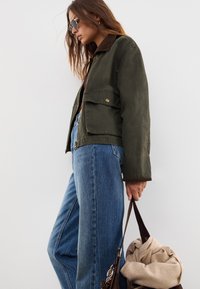 Una donna che indossa occhiali da sole, giacca verde e jeans blu tiene una borsa marrone con un maglione beige contro uno sfondo bianco.
