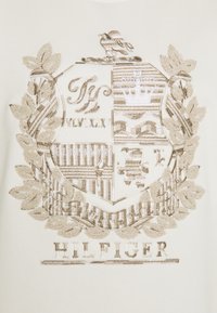 Broderad sköld med intrikata mönster i beige tråd på ett ljust krämfärgat tyg. Innehåller lagerblad och varumärket "HILFIGER" nedanför.