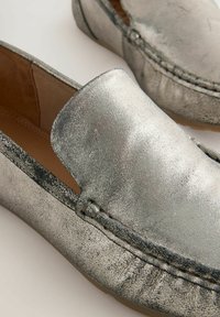 Metalni srebrni loafers s glatkom teksturom, zaobljenim vrhom i ukrasnim šavovima. Mekano unutarnje oblaganje s ravnom potplatom.
