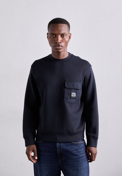 Marineblå crew neck sweatshirt lavet af bomuldsblanding, har en frontlomme med et mærke. Båret med blå jeans, glat tekstur.