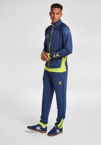 Marinblå träningsoverall med klara gröna accenter; strukturerad axeldetalj, vit chevron på ärmarna och logo på bröstet och byxorna. Mjuk tygblandning.