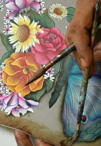 Verfkwasten gehouden boven een gedetailleerd schilderij van bloemen en vlinders. Bevat levendige kleuren: gele zonnebloemen, rode rozen en blauwe accenten.