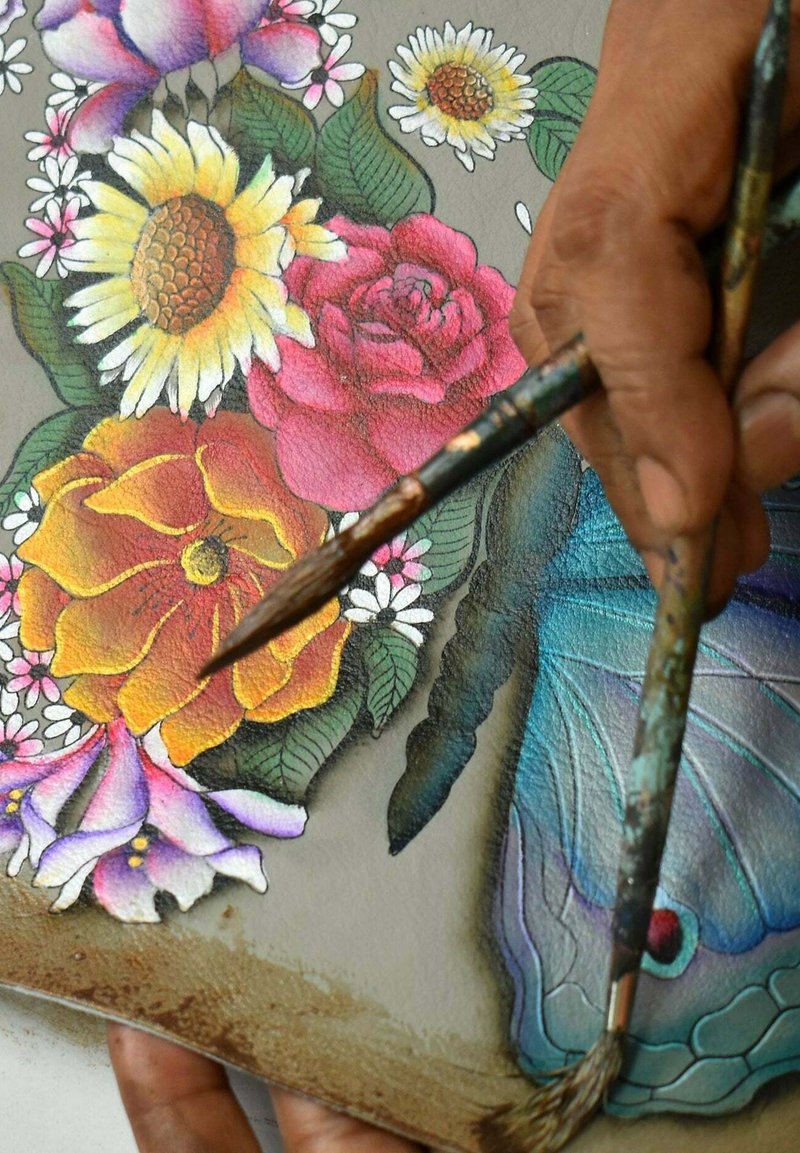 Verfkwasten gehouden boven een gedetailleerd schilderij van bloemen en vlinders. Bevat levendige kleuren: gele zonnebloemen, rode rozen en blauwe accenten.