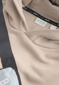 Sweat à capuche beige Ted Baker avec étiquettes de marque visibles et une sangle de sac noire et grise partiellement visible sur une surface sombre.