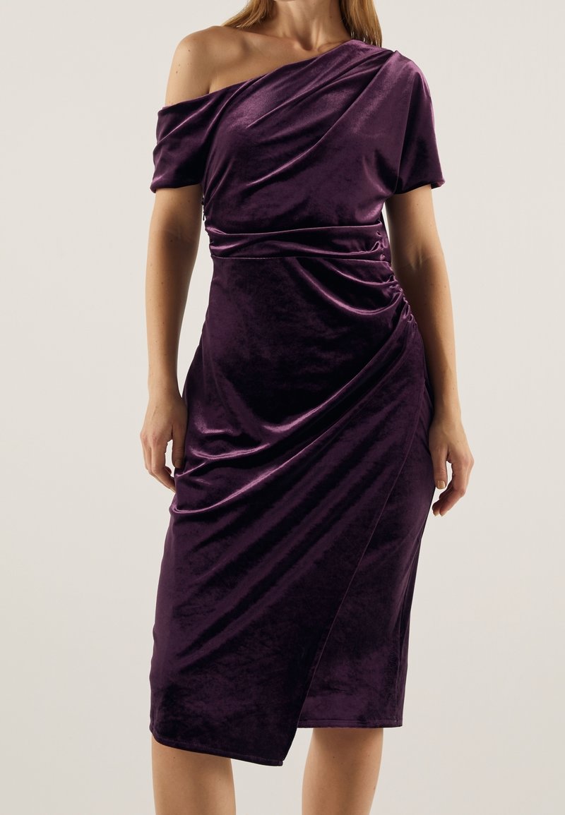 Robe en velours violet profond avec un design asymétrique dénudé d'une épaule, un tissu drapé et une silhouette ajustée atteignant le milieu du mollet.