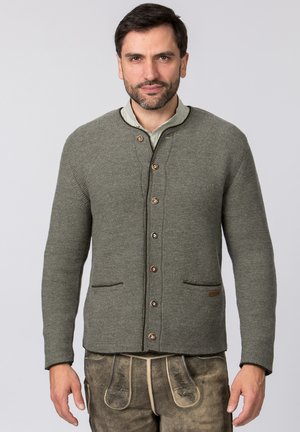 Mann trägt eine graue, gestrickte, geknöpfte Strickjacke über beige traditionelle Lederlederhosen mit Stickerei.