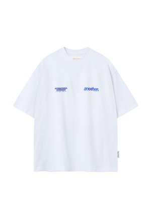 Camiseta blanca oversize con texto azul "AMISTAD INTERNACIONAL a través del deporte" y "another" en el frente, mangas cortas, cuello redondo.