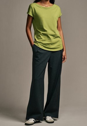 T-shirt vert à manches courtes avec un col rond, associé à un pantalon large et foncé ainsi qu'à des baskets blanches avec des rayures noires.