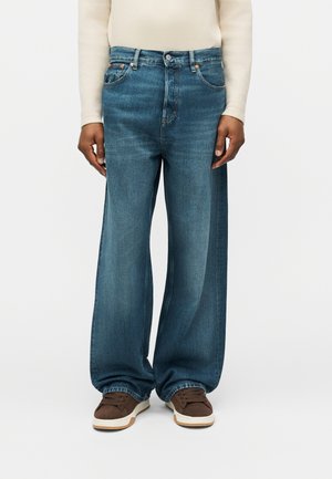 Άτομο που φοράει τζιν παντελόνι με φαρδύ πόδι σε μπλε απόχρωση, λευκή μακρυμάνικη μπλούζα και καφέ αθλητικά παπούτσια, στέκεται μπροστά σε απλό φόντο.