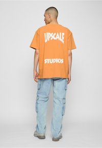 Oranje oversized katoen t-shirt met "UPGRADE STUDIOS" in het wit gedrukt, gecombineerd met lichtblauwe cargojans en grijze sneakers.