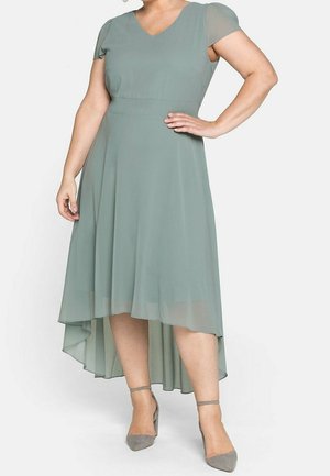 Vrouw in een saliegroene high-low jurk met korte doorzichtige mouwen en grijze enkellaarzen met riempjes, poserend met één hand op haar heup.