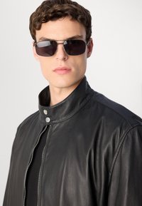 Joven con gafas de sol oscuras y una chaqueta de cuero negra con un cuello alto con cremallera, en un fondo claro y sencillo.