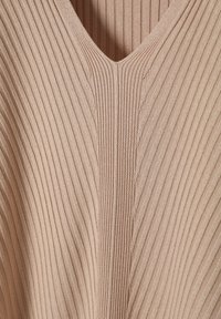 Top de punto acanalado en color beige claro con escote en V. Presenta líneas verticales y una textura suave, destacando la elasticidad y flexibilidad de la tela.