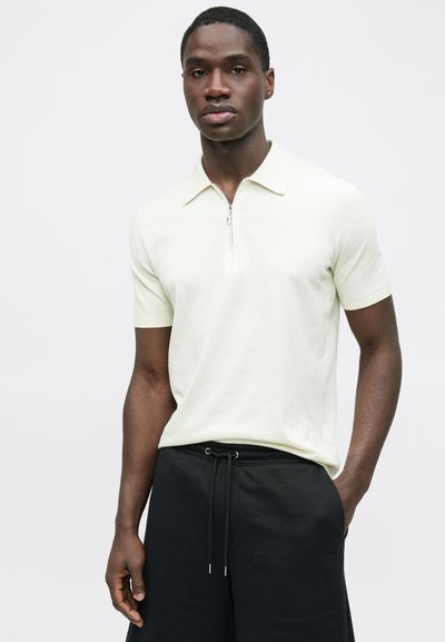 Polo manches courtes jaune pâle, doté d'une fermeture éclair au col et d'une texture lisse, associé à un pantalon noir à cordon de serrage.