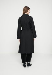 Melns garš tranče mētelis ar jostasvietu, pogotiem apročiem un gludu tekstūru. Valkāts pār vaļējām melnām biksēm un auduma sporta apaviem.