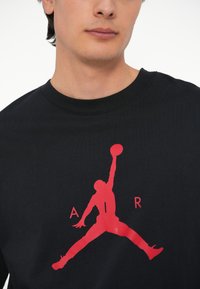Must musta t-särk, millel on suur punane Jumpman logo ja selle all "AIR" tekst. Klassikaline ümar kaeluse disain. Sile tekstuur.