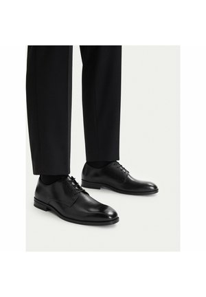 Schwarze, polierte Lederschuhe, getragen mit schwarzen Hosen und dunklen Socken auf hellem Hintergrund.