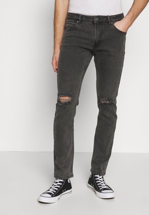 Mężczyzna w czarnych, podartych, obcisłych dżinsach z ręką w kieszeni i czarnych wysokich trampkach, stojący na tle jednolitego białego tła.