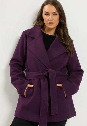 Femme portant un manteau ceinturé violet foncé avec de larges revers, les mains dans les poches, debout devant un mur clair uni.