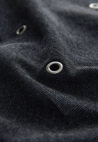Schwarzer Denim-Stoff zeichnet sich durch ein subtil strukturiertes Gewebe aus, auf dessen Oberfläche silberne Ösen sichtbar sind, die Dimension und Detail hinzufügen.