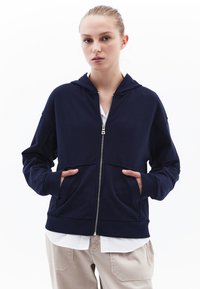 Felpa con zip di colore navy, dotata di tasca frontale, cappuccio e polsini a costine. Realizzata in tessuto morbido, presenta un design pulito e minimalista. Indossata sopra una maglietta bianca.