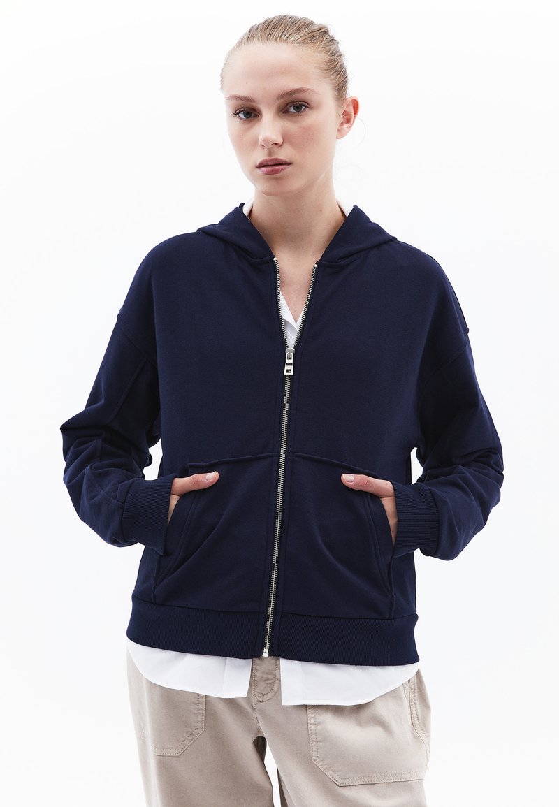 Felpa con zip di colore navy, dotata di tasca frontale, cappuccio e polsini a costine. Realizzata in tessuto morbido, presenta un design pulito e minimalista. Indossata sopra una maglietta bianca.