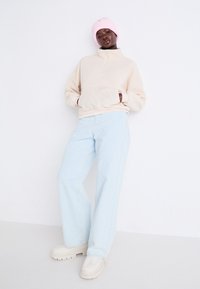 Beige Fleece-Halfzip-Sweatshirt mit einer Vordertasche, kombiniert mit hellblauen weit geschnittenen Jeans und beigen klobigen Schuhen. Rosa Strickmütze.