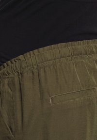 Zielona oliwkowa talia z elastycznym paskiem i płaskim detalem kieszeni, połączona z czarnym materiałem powyżej. Miękka tekstura, casualowy design.