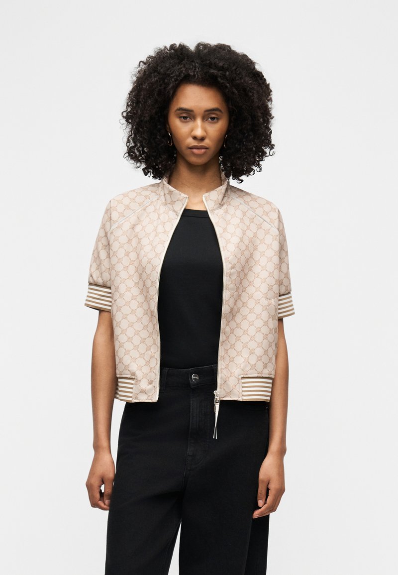 Γυναίκα με κατσαρά μαλλιά, που φοράει μπεζ μπουφάν με φερμουάρ και σχέδια, με κοντά μανίκια, πάνω από μαύρο τοπ και μαύρο παντελόνι, στέκεται μπροστά σε απλό φόντο.