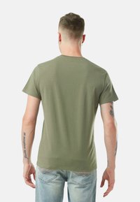 Homme aux cheveux courts portant un t-shirt vert olive et un jean bleu clair, de dos, montrant des tatouages sur les deux bras et des boucles d'oreilles.