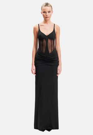 Femme aux cheveux blonds dans une élégante robe de soirée noire avec un corsage transparent et une taille froncée, debout devant un fond blanc uni.