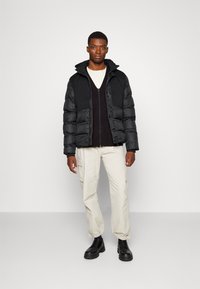 Sort pufferjakke med hætte, grå strikket sweater under, cream cargo bukser og sorte snørede støvler. Står på en ensfarvet baggrund.