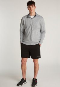 Joven de pie en pantalones cortos negros, chaqueta gris con cremallera y zapatillas negras contra un fondo claro y liso.