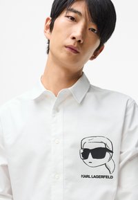 Baltas marškinėliai su sagomis, ant kurių siuvinėtas juodas piešinys žmogaus su akinių nuo saulės ir tekstas "KARL LAGERFELD" žemiau.