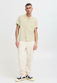 Chemise verte claire à manches courtes avec une poche poitrine, bouton devant, et une coupe décontractée, associée à un pantalon blanc cassé et des baskets noires.