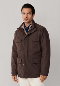 Hnedá bunda s viacerými vreckami, s golierom, látka pripomínajúca tvíd, kontrastný vnútorný golier a zapínanie na zips vpredu.