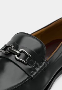 Mocassin en cuir noir à finition lisse et brillante. Comprend un accent en chaîne métallique et des détails cousus le long du bord supérieur. Doublure intérieure marron.