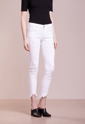 Femme portant un jean skinny blanc déchiré, un top noir à manches courtes et des mules noires à talons, debout sur un sol beige.