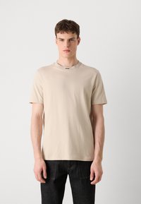 T-shirt beige à manches courtes en coton lisse, avec un col rond à bord côtelé et un petit logo noir sur le col.