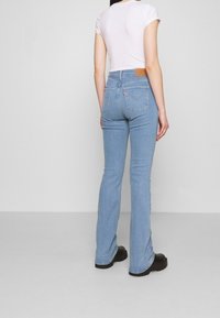 Světle modré zvonové džíny vyrobené z denim materiálu, s koženou nášivkou na zadní straně a tradičními zadními kapsami. Nosené s bílým tričkem s krátkým rukávem.