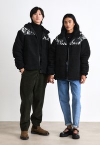 Black fleece jackets with patterned shoulder accents, zippered front, worn with dark green corduroy pants and light blue jeans. Black footwear.