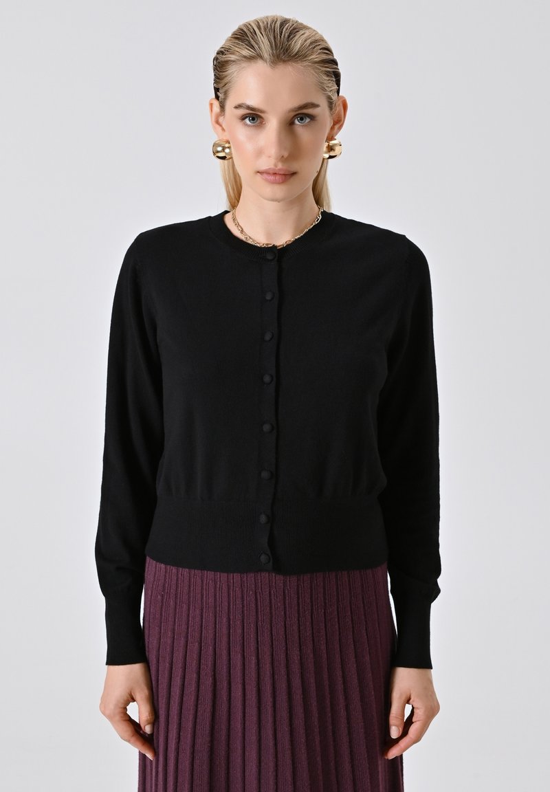 Frau in einem schwarzen, knopfartigen Cardigan, mit großen goldenen Ohrringen und einem lila, plissierten Rock, die vor einem schlichten, hellen Hintergrund steht.