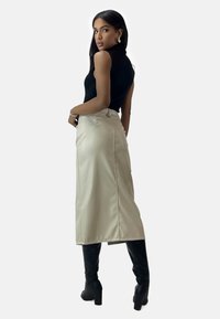 Falda midi de color beige claro con una textura suave, abertura trasera y bolsillos laterales, combinada con una blusa ajustada negra sin mangas y botas negras hasta la rodilla.