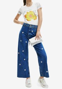 White ribbed t-shirt with a yellow flower graphic. Blue wide-leg denim jeans with white daisy embellishments and silver clutch bag.