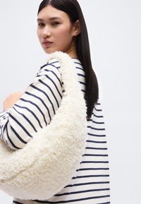 Νέα γυναίκα με μακριά σκούρα μαλλιά που φοράει λευκή μπλούζα με ναυτικές ρίγες, κρατώντας μια μεγάλη, αφράτη λευκή τσάντα ώμου, κοιτάζοντας από πάνω από τον ώμο της.