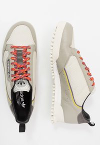 Beige idrottsskor med övre del av mesh och läder, färgglada mönstrade snören, texturerad sula och påtagliga gula och lila accenter.