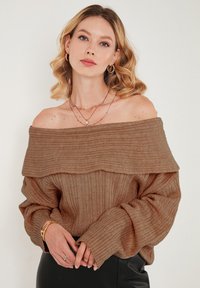 Suéter marrón de un solo hombro con textura acanalada, mangas largas y detalle de cuello doblado. Combinado con collares dorados en capas y un pantalón negro.