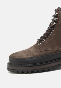 Stivale alla caviglia in suede marrone con intersuola in gomma nera, occhielli in metallo, lacci grigi e suola strutturata e scolpita, evidenziando durabilità e aderenza.