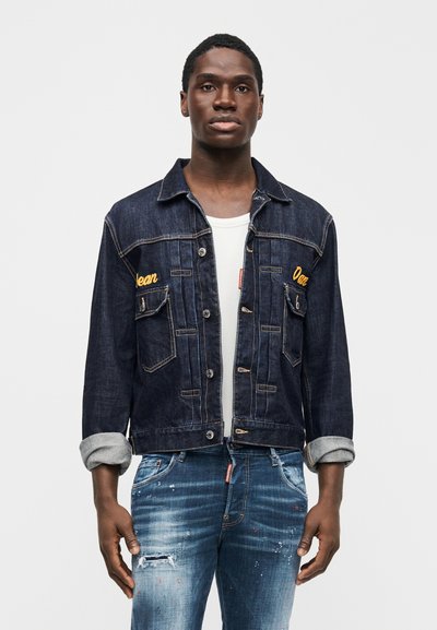 Hombre con chaqueta de mezclilla oscura con bordado amarillo sobre camisa blanca y jeans azules desgastados contra fondo blanco liso.