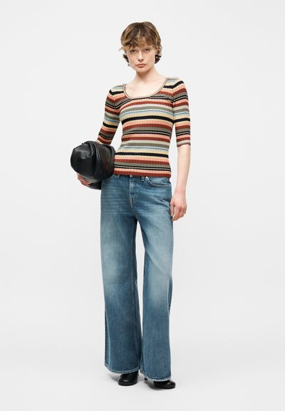 Άτομο που φοράει ριγέ κοντομάνικη μπλούζα και φαρδιά μπλε τζιν κρατώντας μια μαύρη τσάντα μπροστά σε απλό φόντο.